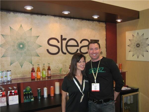 A dark-haired woman and a man in a black Steaz shirt stand smiling in front of a Steaz logo wall, with bottles and cans displayed on shelves behind them.