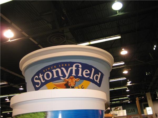 A large display shaped like a Stonyfield yogurt tub, with a cow illustration and the text Since 1983, hangs under the ceiling lights of the exhibit hall.