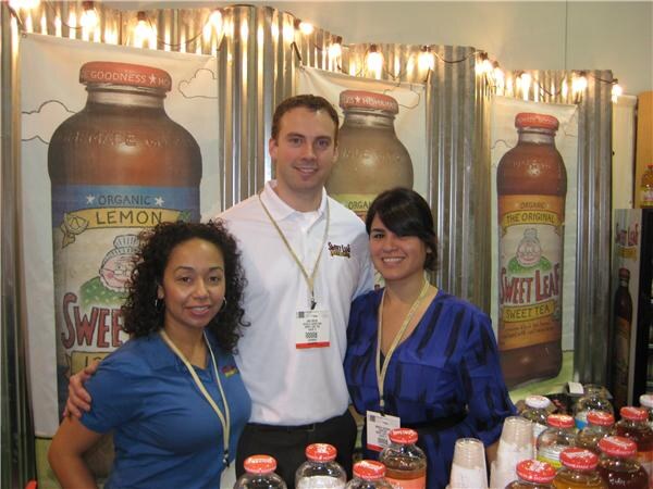 Two women and a man in a white polo smile for the camera at the Sweet Leaf booth, with large Sweet Leaf bottle graphics behind them and bottles on the table in front.