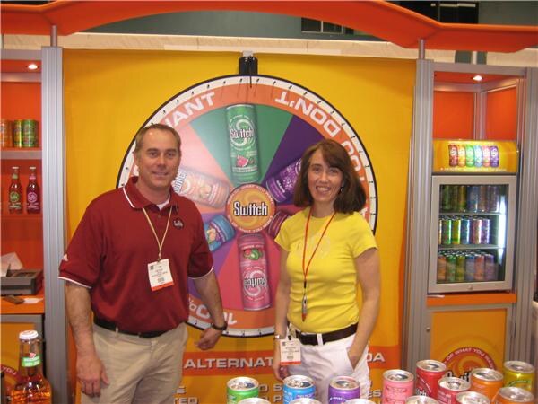 A man in a maroon polo and a woman in a yellow shirt stand in front of a Switch soda booth with a prize wheel showing Switch cans; soda cans are in the foreground.