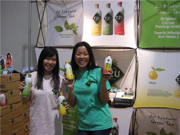 Two smiling women hold bottles of T2U green tea in front of T2U banners reading All Natural Green Tea, with stacked cardboard boxes to the left.