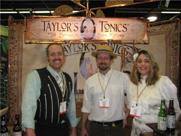 Two men and a woman in vintage-style clothing stand beneath a Taylor's Tonics The Botanical Soda Kitchen sign, with bottles on the counter in front of them.