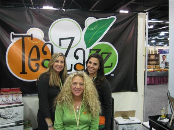 Three smiling women, one seated in front in a green top and two standing behind in black, pose in front of a large orange and green TeaZazz banner.