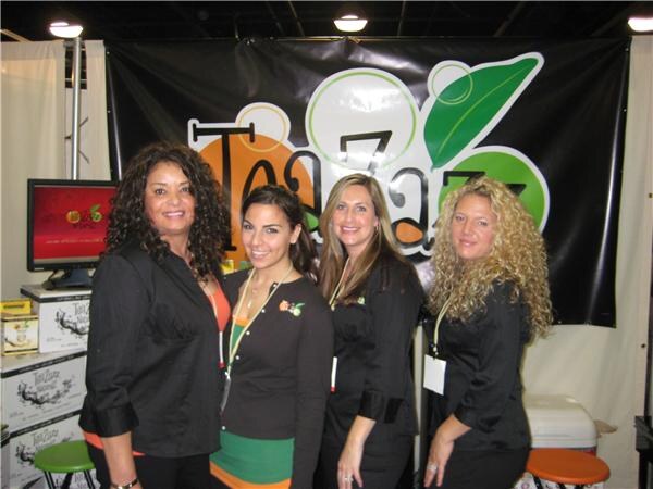 Four women, most dressed in black, stand side by side in front of a black TeaZazz banner, with product boxes and a monitor to the left.
