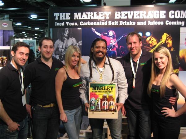 Six people in black shirts pose at The Marley Beverage Company booth. The man in the center holds a wooden box of Marley Beverage Company drinks, with concert photos behind them.