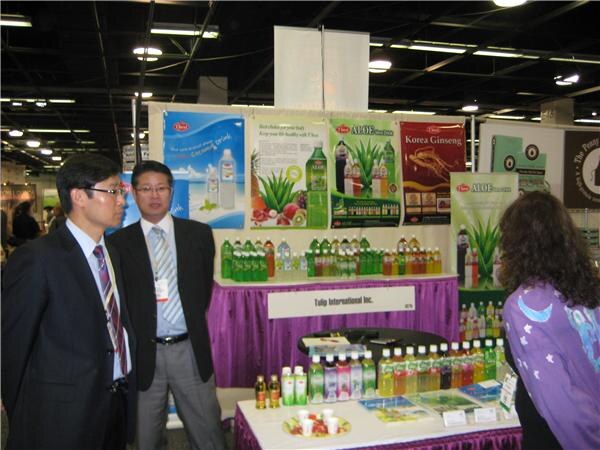 Two men in dark suits talk with a woman in a purple top at the Tulip International Inc. booth, with aloe and Korean ginseng drink posters and bottles on display.