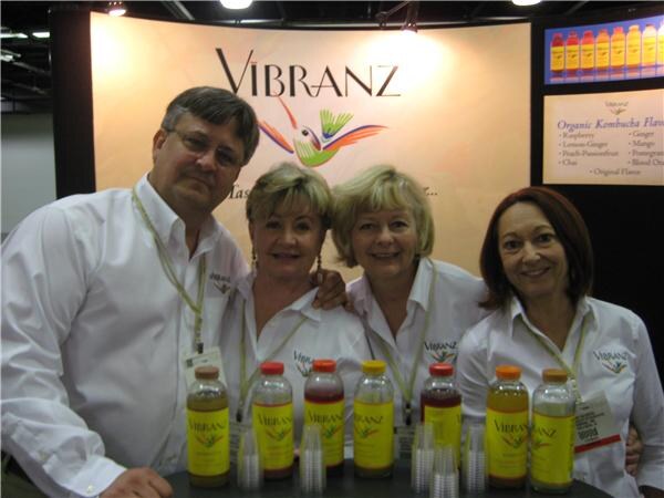 A man and three women in white shirts pose behind a table of Vibranz kombucha bottles in front of a Vibranz sign with a hummingbird logo.