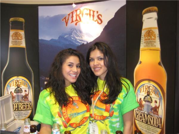 Two smiling women in tie-dye Reed's shirts stand between large cutout Virgil's bottles labeled Root Beer and Cream Soda, with a mountain image behind them.