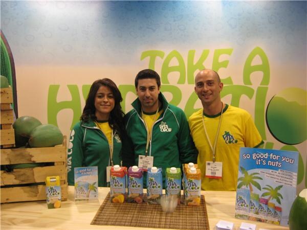Two men and a woman in green and yellow Vita Coco apparel stand behind a counter of Vita Coco cartons, with a "Take a Hydration" backdrop and coconuts in a crate.