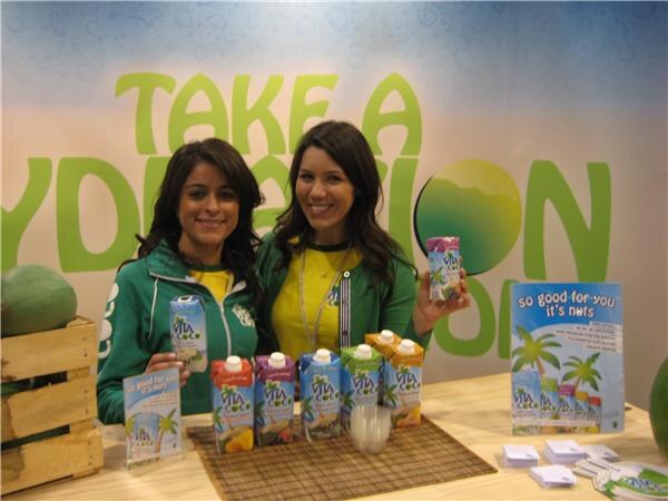 Two smiling women in green jackets hold Vita Coco cartons behind a counter displaying more cartons, in front of a green "Take a Hydration" backdrop.