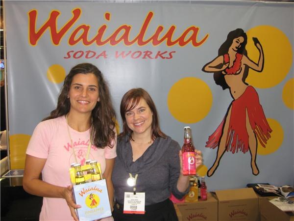 Two women stand in front of a Waialua Soda Works banner with a hula dancer illustration; one holds a four-pack of pineapple soda and the other holds a bottle.