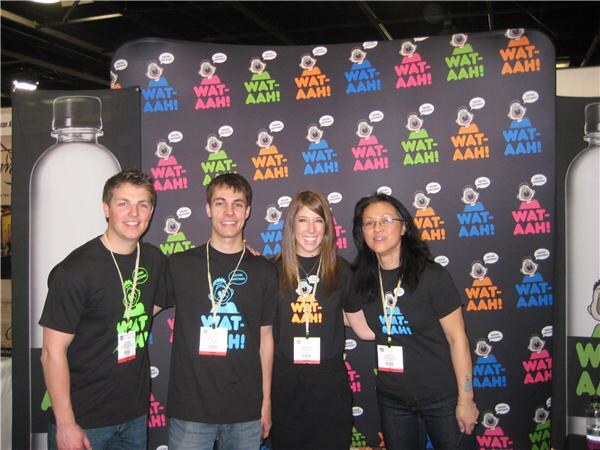 Two men and two women in black Wat-aah! T-shirts stand together in front of a backdrop of colorful Wat-aah! cartoon characters, with large bottle graphics at the sides.