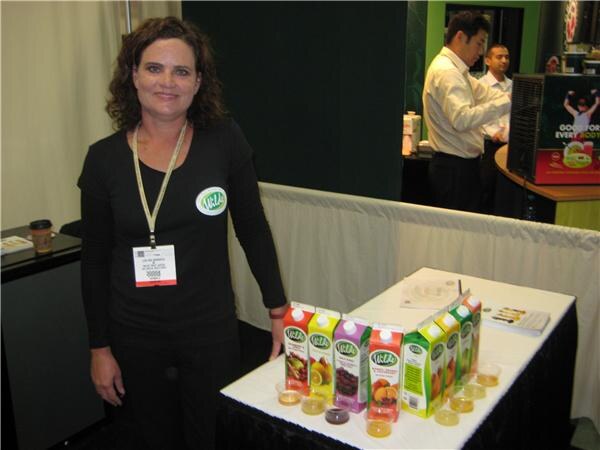 A woman in black with a badge stands beside a table of Wilde juice cartons and small sample cups, with two men talking in the background.
