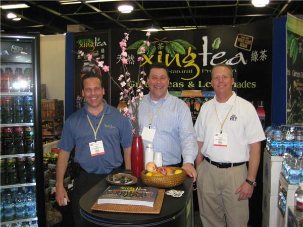 Three men in polo and button-up shirts stand around a table with a fruit bowl and a vase of blossoms in front of a Xing Tea sign.