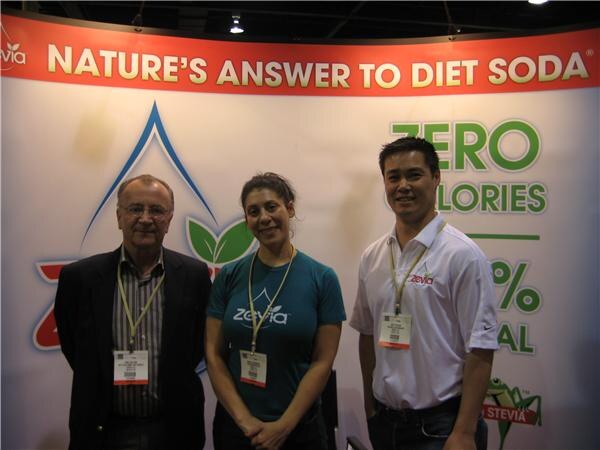Two men and a woman wearing badges stand in front of a Zevia banner reading "Nature's answer to diet soda" and "Zero calories". The woman wears a teal Zevia shirt.