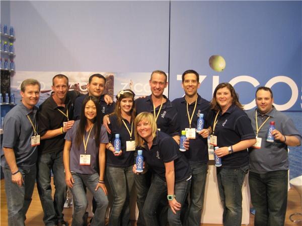 Ten people in navy and gray shirts and badges pose close together at the Zico booth, several holding Zico bottles, in front of a large Zico sign.