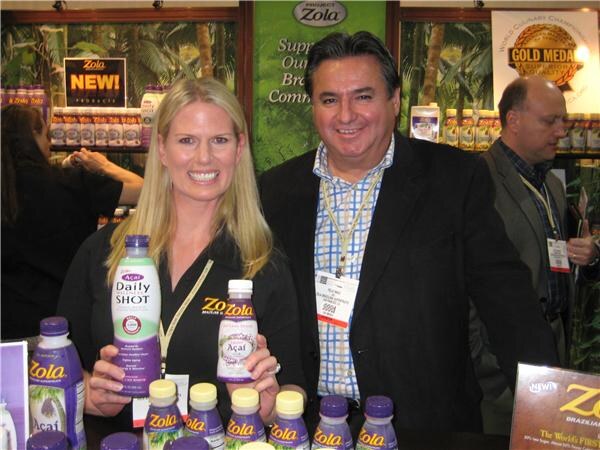 A smiling blonde woman holding Zola acai bottles stands beside a smiling man wearing a badge, with shelves of Zola products and a Project Zola sign behind them.