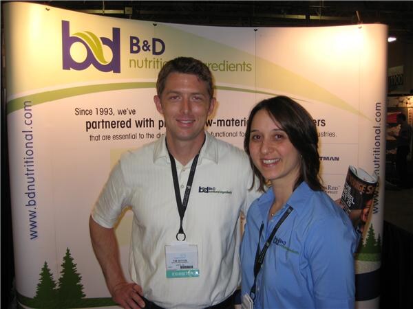 A man in a white striped shirt with an exhibitor badge and a woman in a blue shirt pose in front of a B&D Nutritional Ingredients display.