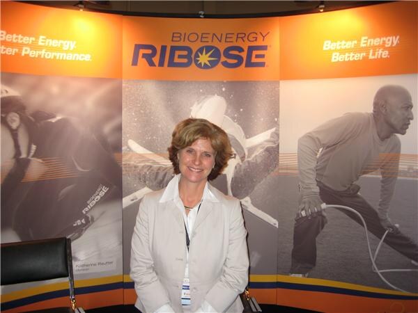 A smiling woman with short brown hair in a light jacket stands in front of BioEnergy Ribose banners reading "Better Energy. Better Performance."