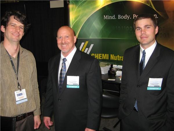 Three men stand in front of a CHEMI Nutra sign; two wear suits and ties with exhibitor badges, and one wears a striped shirt with a lanyard.