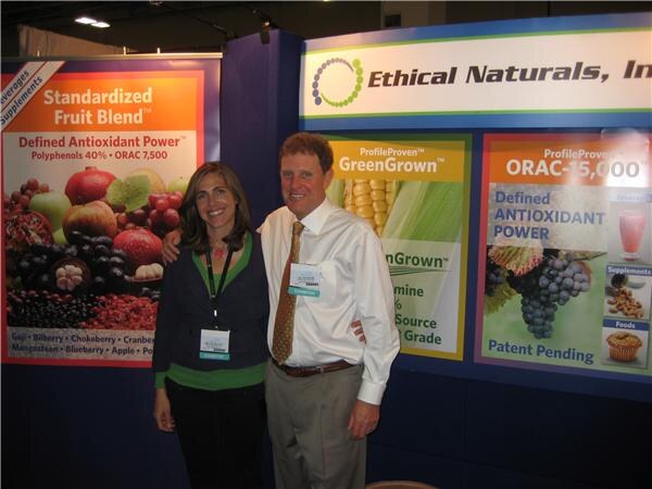 A woman and a man stand close together in front of Ethical Naturals display panels showing fruit, corn and grapes with antioxidant claims.
