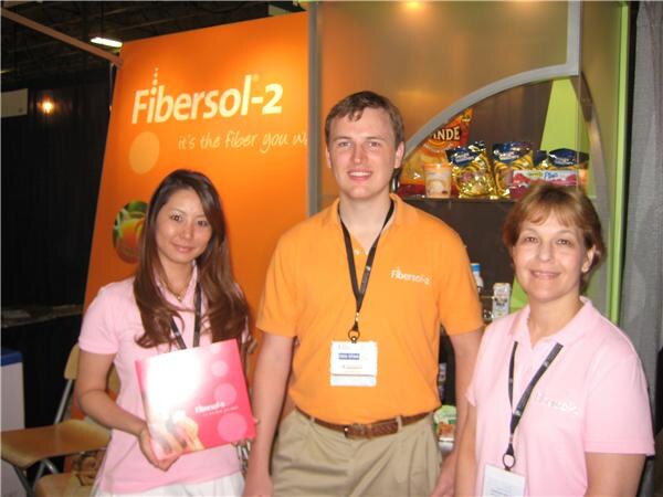 Two women and a man in orange and pink polo shirts stand at a Fibersol-2 booth; one woman holds a pink brochure and snack bags sit on shelves behind them.