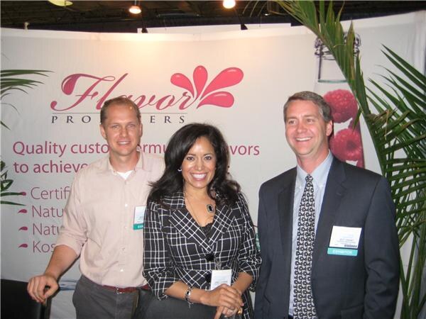 A woman in a black and white checked jacket stands between two men in front of a Flavor Producers banner, with a plant and raspberry graphic nearby.
