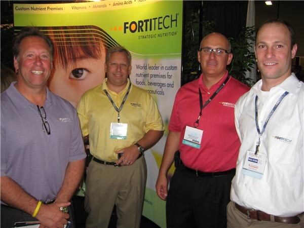 Four men wearing badges and lanyards stand together in front of a green Fortitech banner for custom nutrient premixes.
