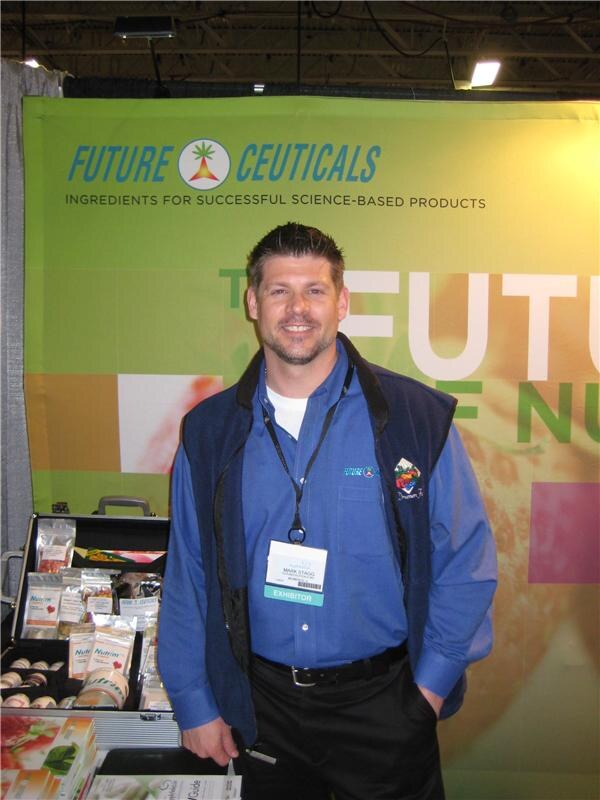 Mark Stagg of Futureceuticals, smiling in a blue shirt and vest with an exhibitor badge, stands in front of a green Future Ceuticals banner beside a table of product packages.