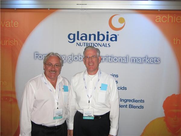 Two smiling men in white shirts with exhibitor badges and lanyards stand in front of an orange and white Glanbia Nutritionals backdrop.