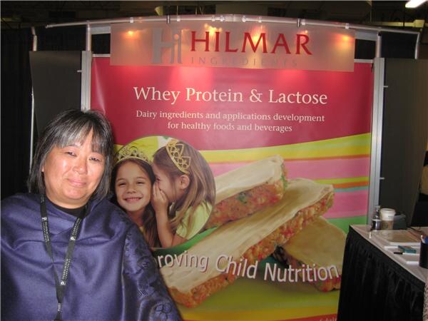 A woman with dark hair and a lanyard stands in front of a pink Hilmar banner reading Whey Protein & Lactose, showing two children and bars.