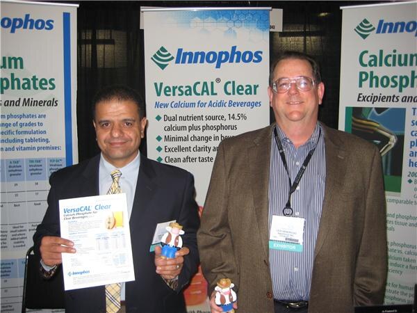 Two men stand before Innophos banners for VersaCAL Clear; the man on the left holds a product sheet and a small figurine, and the man on the right wears an exhibitor badge.
