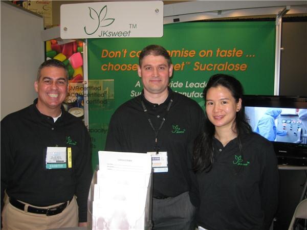 Two men and a woman in black JKsweet polo shirts stand together in front of a green JKsweet sucralose display; two men wear exhibitor badges.