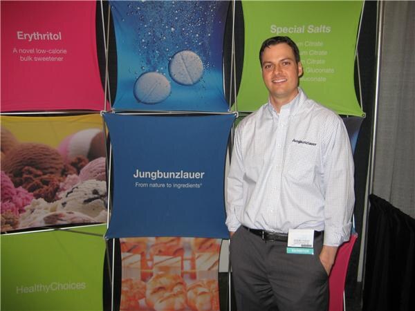 A smiling man in a white checked shirt with an exhibitor badge stands beside Jungbunzlauer display panels showing erythritol, ice cream and special salts.
