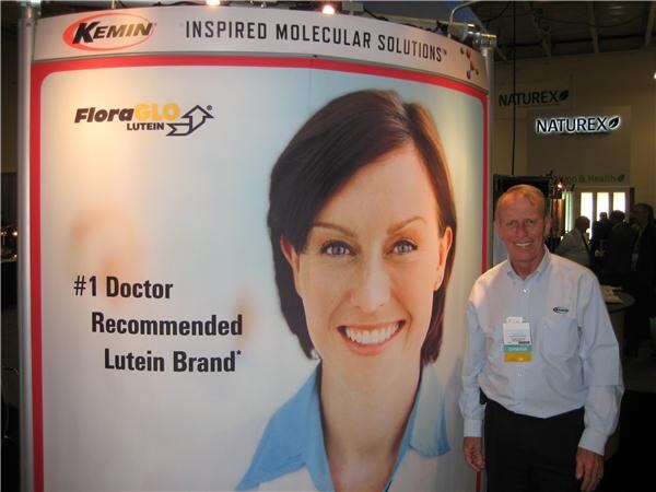 A smiling older man in a light shirt stands beside a Kemin FloraGLO Lutein display with a large woman's portrait; a Naturex sign is visible in the background.