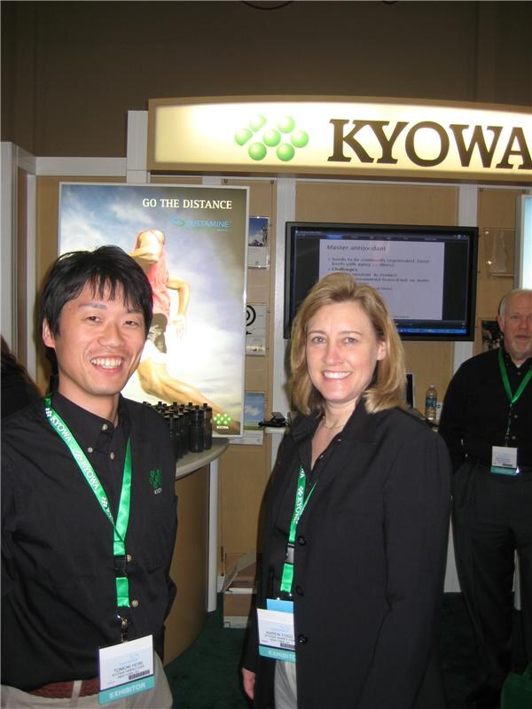 Tomoki Hori of Kyowa, left, and a woman in a black jacket, both smiling and wearing green lanyards, stand before a Kyowa booth with a Sustamine poster and a monitor.