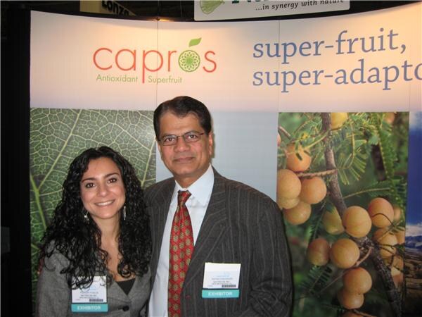 A woman with dark curly hair and a man in glasses and a red tie, both wearing exhibitor badges, stand in front of a Capros antioxidant superfruit banner.