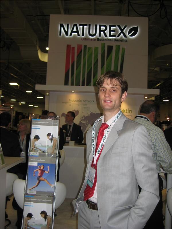 A man in a gray suit and red tie with a Naturex lanyard stands in front of the lit Naturex sign and a brochure rack on a busy exhibit floor.