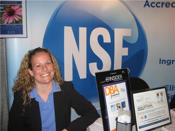 A smiling woman with curly hair in a black blazer and blue shirt sits in front of a large NSF logo. A DBA Analytical display and a framed sign rest on the table beside her.