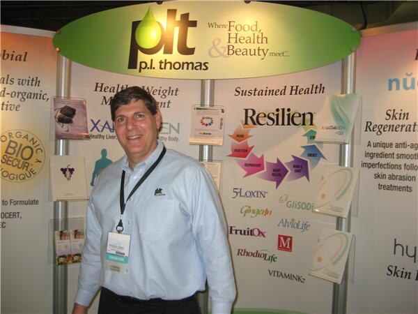 A smiling man in a light blue shirt with an exhibitor badge stands in front of a p.l. thomas display listing Resilien, 5-Loxin, FruitOx, RhodioLife and Vitamink.