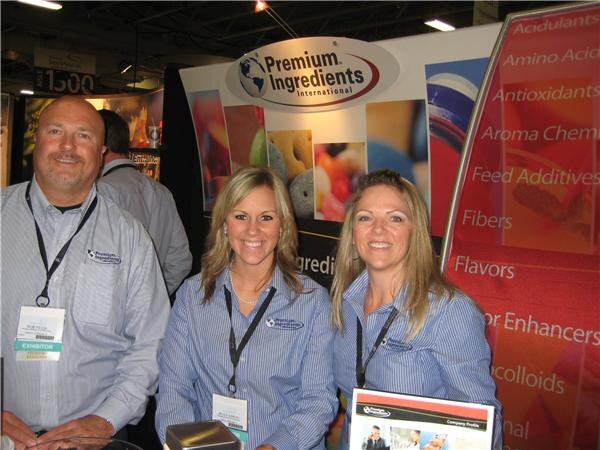 A man and two women in matching striped shirts and exhibitor lanyards smile in the Premium Ingredients International booth, with a red banner listing ingredient categories behind them.