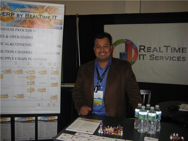 A smiling man in a brown blazer and blue shirt stands behind a table with water bottles, candy and brochures. A RealTime IT Services banner hangs behind him.