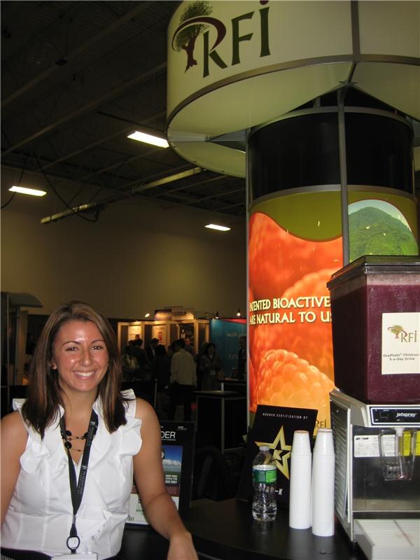 A smiling woman in a white sleeveless top with a lanyard stands beside an RFI display tower. A Jetspray drink dispenser, stacked cups and a water bottle are on the counter.