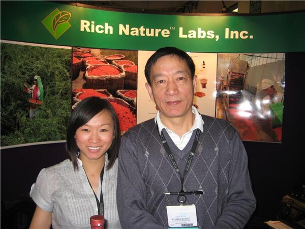 Richard Zhang, in a gray argyle sweater and badge, stands beside a smiling woman in a gray top. A Rich Nature Labs, Inc. sign and photos of red berries are behind them.