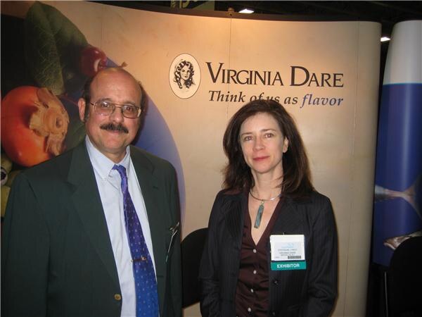 A mustached man with glasses in a green blazer and purple tie stands beside a woman in a dark jacket with an exhibitor badge, in front of a Virginia Dare sign.