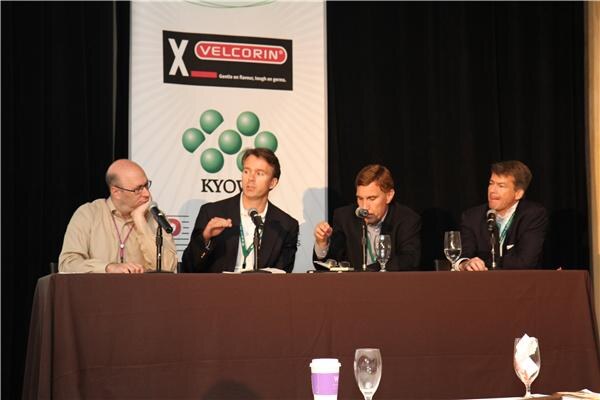 Four men sit at a draped table with microphones during a panel; a banner behind them shows the Velcorin logo and partial green logo text.