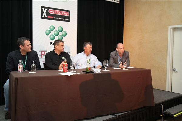 Four men sit at a table with a brown tablecloth, with water glasses and bottles in front of them, beside a Velcorin banner on a stage.