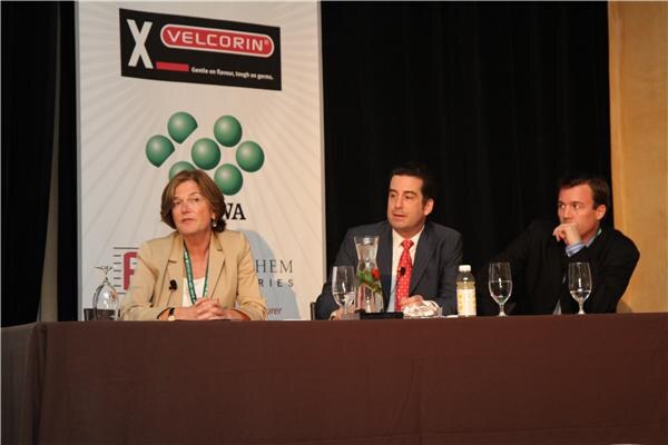 A woman in a beige blazer and two men in dark jackets sit at a table with water glasses and a carafe, beside a Velcorin banner.