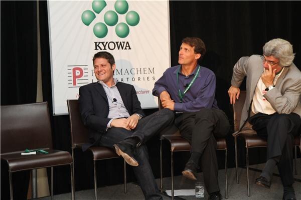 Two men in jackets and one older man with gray hair sit in chairs on a stage in front of a banner reading Kyowa and Pharmachem Laboratories.