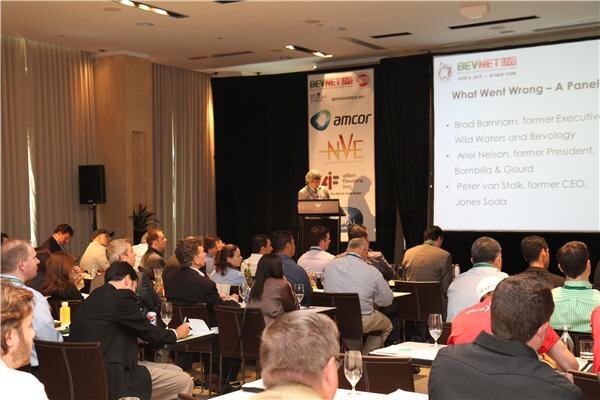 An audience seated at tables faces a podium and a projection screen reading "What Went Wrong – A Panel," beside a banner for Amcor and NVE Pharmaceuticals.